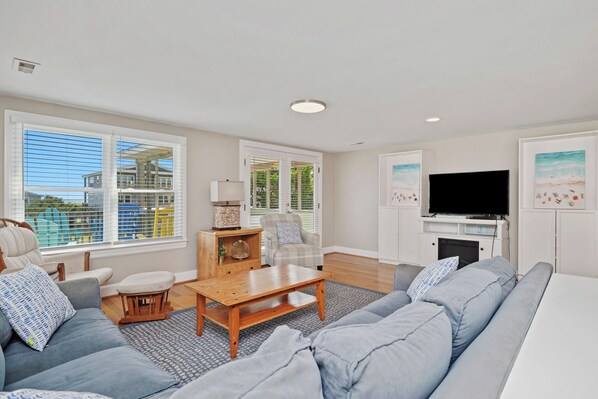A spacious living room with a large sofa, a wooden coffee table, and a television. There are large windows with blinds, a fireplace, and a view of buildings outside.