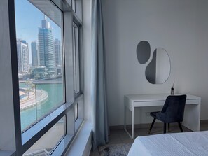 Ein modernes Hotelzimmer mit großem Fenster und Blick auf die Stadt, einem weißen Schreibtisch, einem schwarzen Stuhl und einem runden Spiegel an der Wand.