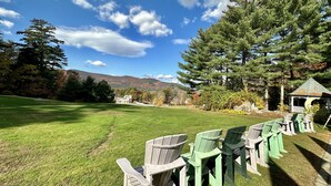 Un espace extérieur verdoyant, avec des chaises Adirondack disposées en rangées, un belvédère et un paysage montagneux en toile de fond, sous un ciel partiellement nuageux.