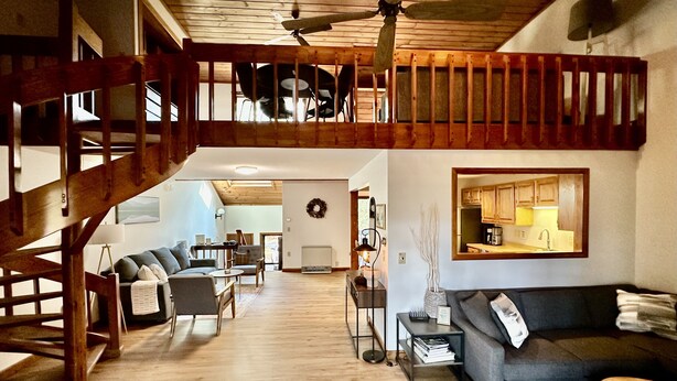 Un vaste espace de vie doté d’un haut plafond, d’un escalier en bois et d’une cuisine visible à travers une porte ouverte.