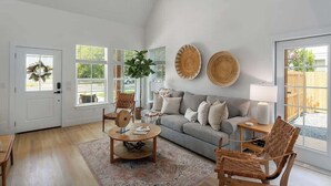 Un salon spacieux avec un canapé gris, une table basse en bois et des fauteuils en osier. Il y a deux grandes fenêtres à cadres blancs, une porte ornée d’une guirlande et une lampe fixée au mur.