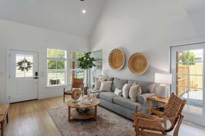 A spacious living room with a grey sofa, wooden coffee table, and wicker chairs. There are two large windows with white frames, a door with a wreath, and a wall-mounted lamp.