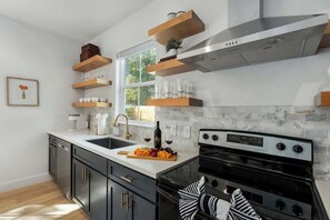 A modern kitchen with dark cabinets, a white countertop, a stainless steel range hood, and wooden shelves above the sink.