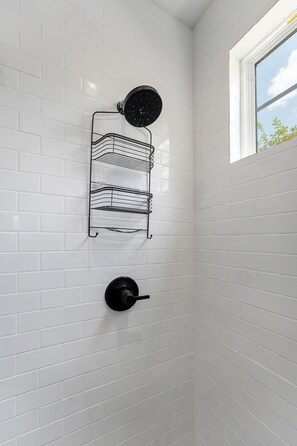 A bathroom with white tiled walls, a black showerhead, and a black towel rack. - The Noir: Hot-tub + Firepit 3 Blks to Main Street! (Fredericksburg)
