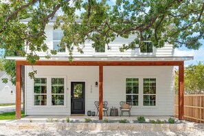 A white house with a covered porch, two windows, a door, and a small seating area with chairs and a table. - The Noir: Hot-tub + Firepit 3 Blks to Main Street! (Fredericksburg)