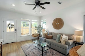 Un salon spacieux avec un canapé gris, une table basse en verre, un ventilateur de plafond et une grande fenêtre donnant sur un paysage verdoyant.
