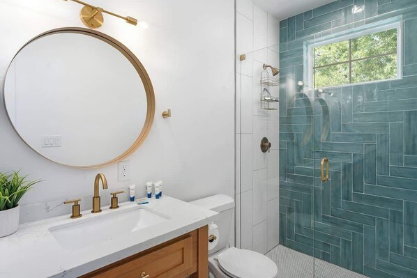 A bathroom with a white sink, gold faucets, a round mirror, and a blue-tiled shower enclosure.