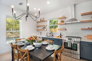 A modern kitchen with a dining table set for six, wooden chairs, a chandelier, built-in shelves, and a stainless steel oven. - The Malbec: 3 Blocks to Main St w/Hot-Tub+Firepit! (Fredericksburg)