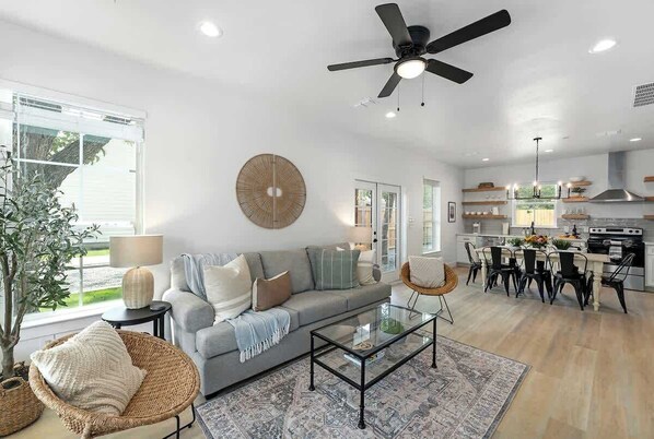 A modern living room with a grey sofa, a glass coffee table, and a dining area with a kitchen in the background.