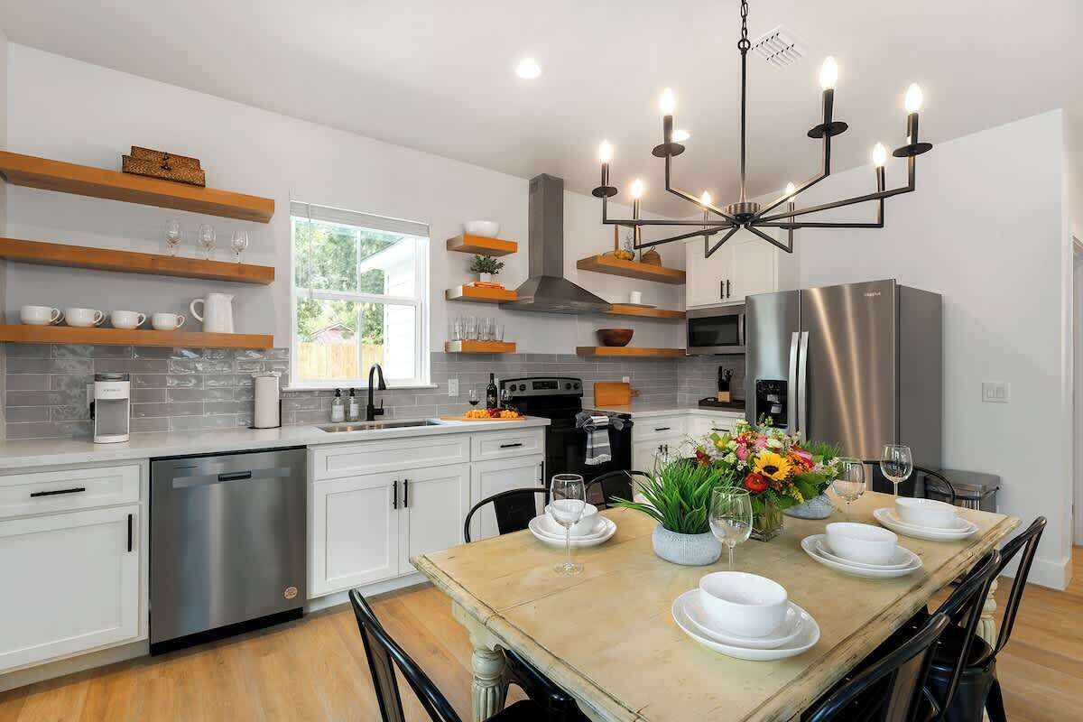 A modern kitchen with white cabinets, a stainless steel dishwasher, a built-in oven, and a large refrigerator. There is a dining table set with plates and glasses, and a chandelier hangs above.