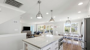 Una cocina moderna con una isla de mármol, armarios blancos y electrodomésticos de acero inoxidable. Hay un comedor con sillas blancas y una mesa, y una sala de estar con un sofá y una televisión.