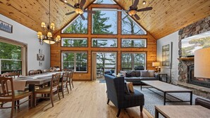 A spacious room with a high ceiling, large windows, a fireplace, and a dining area. - Lakeside: 4 Kings, Room4Groups, Hot Tub, Dock, Firewood (Brainerd)