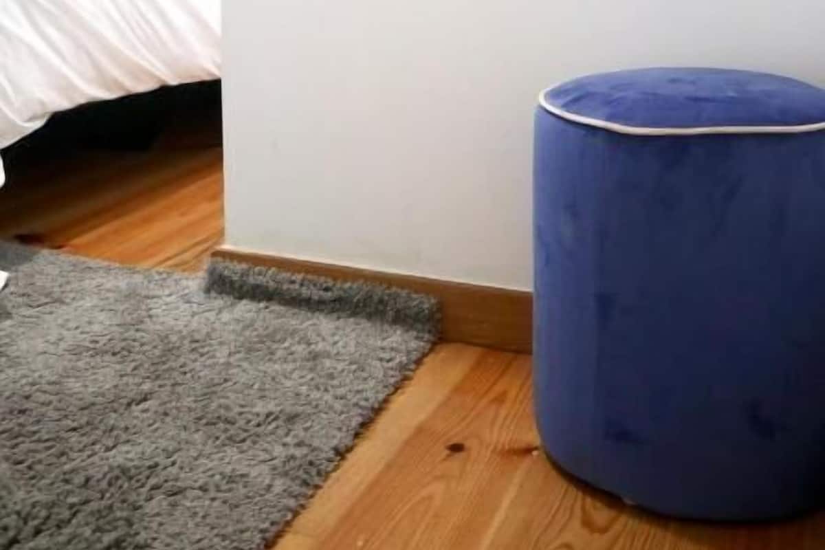 Une chambre à coucher avec un lit, un grand miroir, un pouf bleu et un tapis gris.