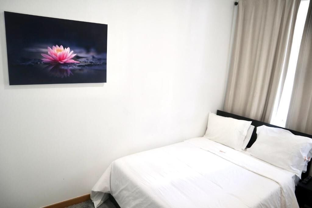 Une chambre d’hôtel avec un lit, du linge de lit blanc, un tableau représentant une fleur de lotus rose et une fenêtre avec des rideaux.