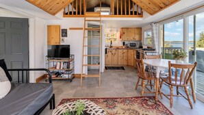 Un living in stile loft con soffitto in legno, un letto a soppalco, una cucina con mobili in legno e un'area pranzo con sedie in legno.