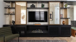 A modern living room with a large flat-screen TV mounted on a marble wall, a built-in fireplace, and a black entertainment unit with shelves and decorative items. - HIGHSTAY - Luxury Serviced Apartments - Tuileries Garden (Paris)