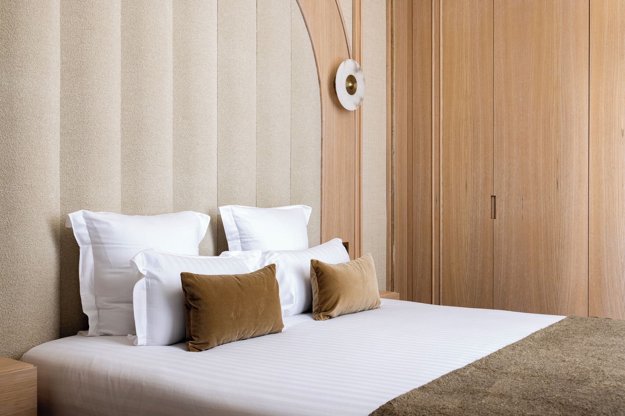 A neatly made bed with white and brown pillows, a wooden headboard, and a textured wall behind.