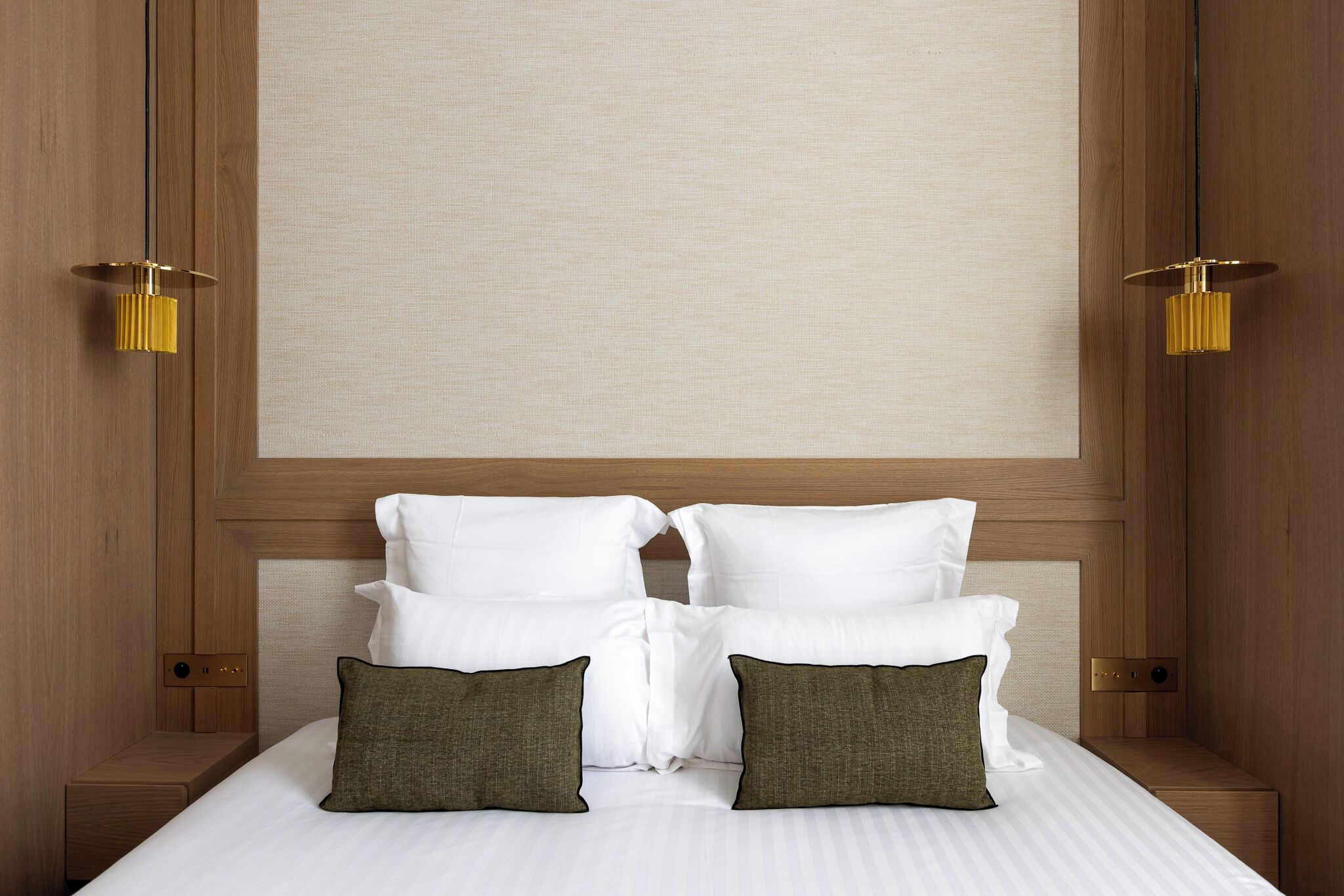 A neatly made bed with white linens and two green pillows, a wooden headboard, and two wall-mounted lamps.