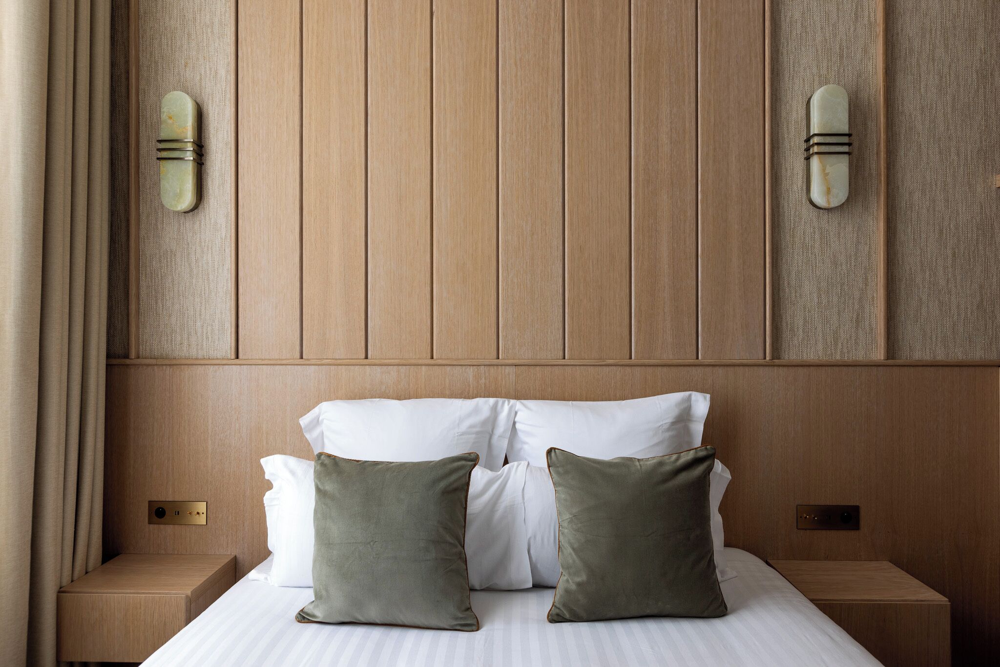 Cama bien hecha con sábanas blancas y dos almohadas verdes, cabecero de madera y mesitas de noche con lámparas.