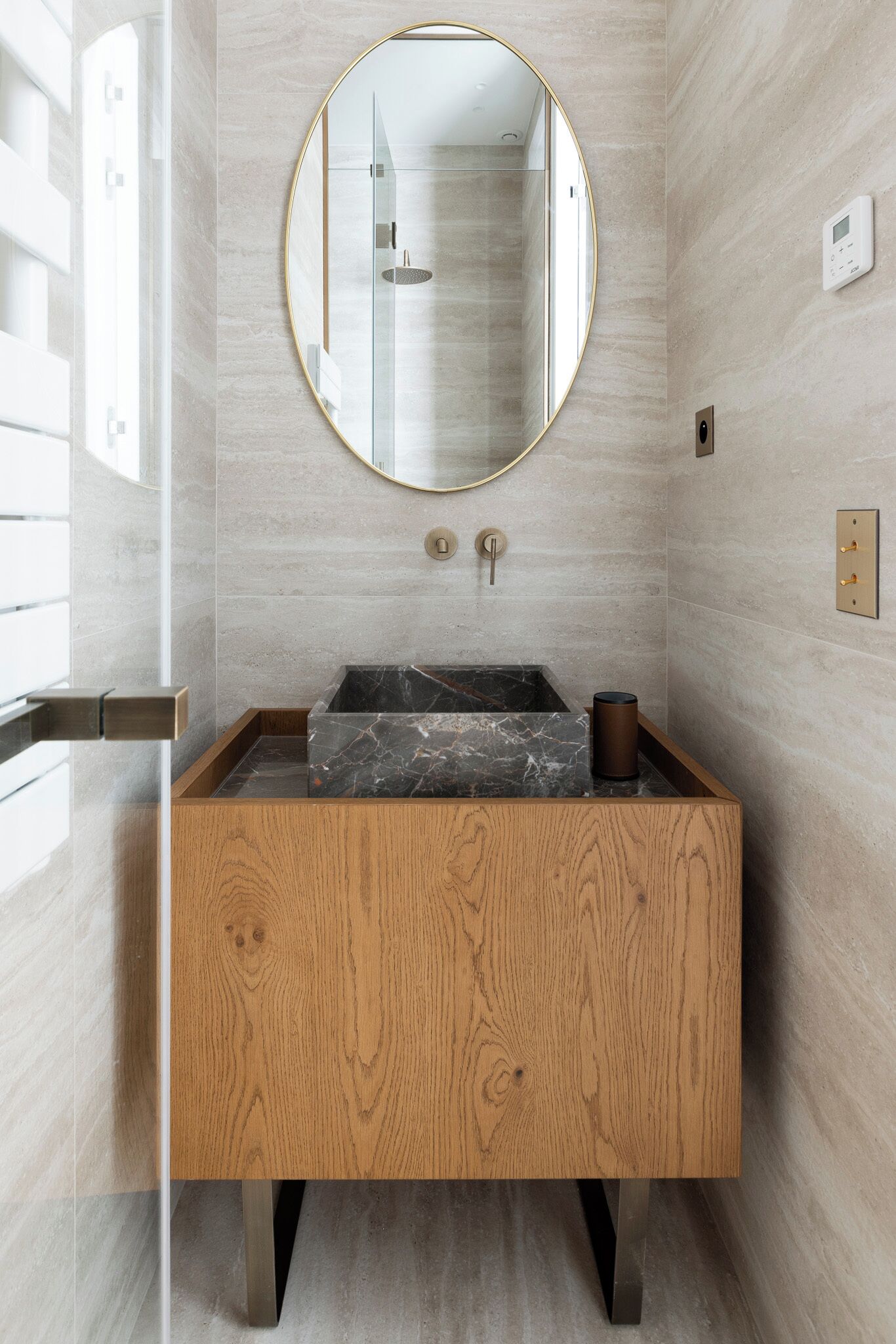 A bathroom with a wooden vanity, a marble sink, a round mirror, and a glass shower enclosure.