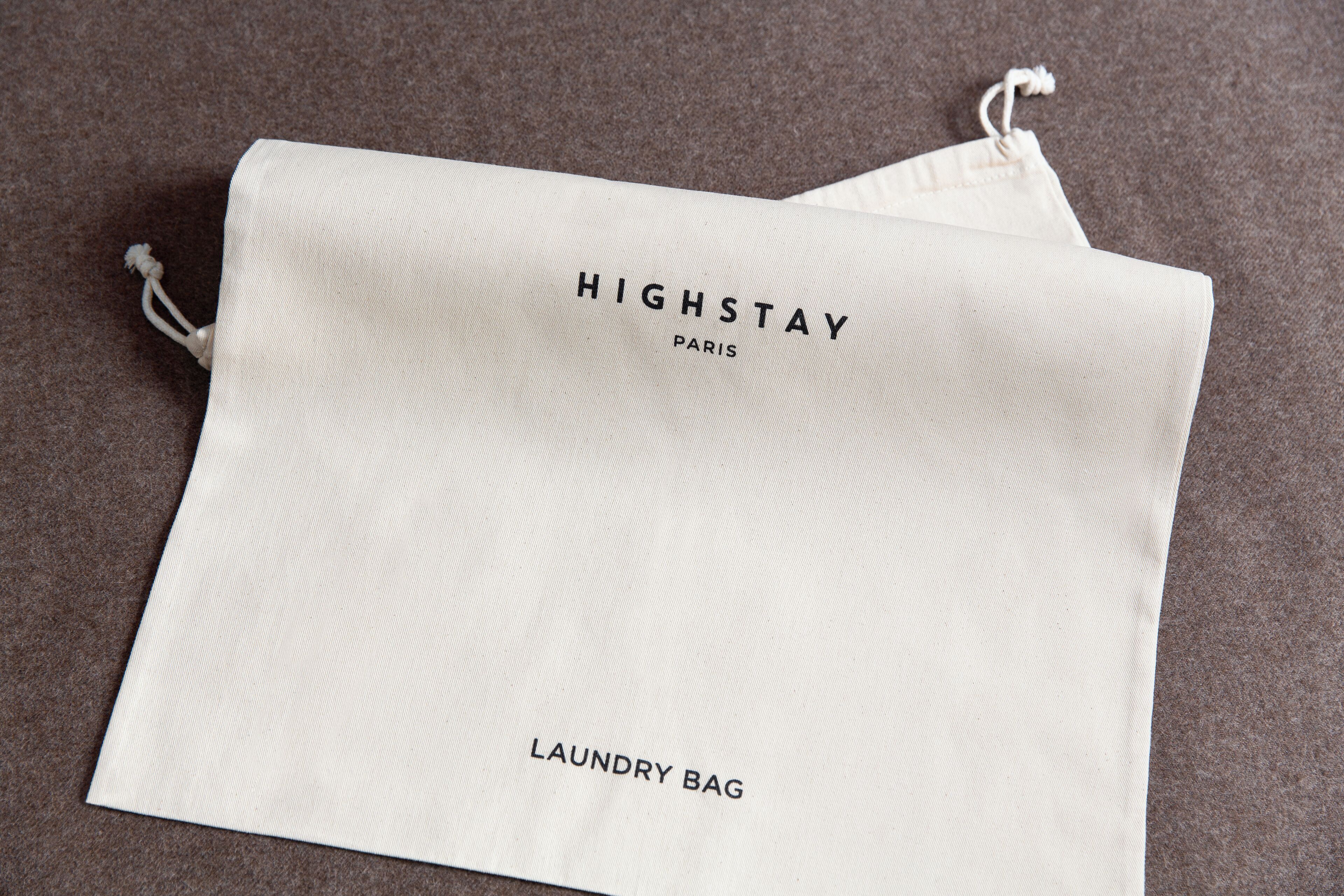 Una bolsa blanca con cordón de tiro, etiquetada como "HIGHSTAY PARIS LAUNDRY BAG", sobre una superficie texturizada.