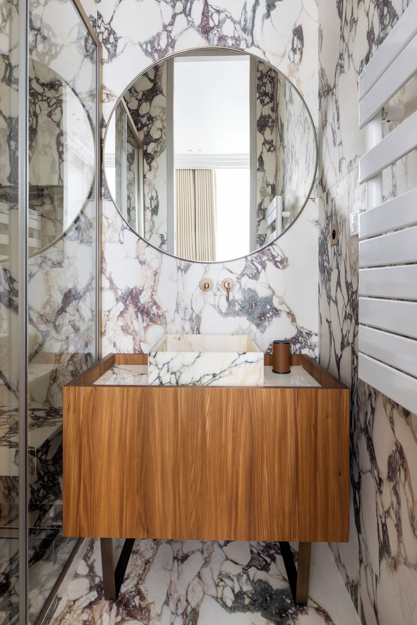 A bathroom with a marble wall, a wooden vanity, a round mirror, and a towel rack.