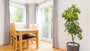 A dining area with a wooden table and chairs, a potted plant, and large windows with curtains. - Edinburgh Aparthotel (Edinburgh)