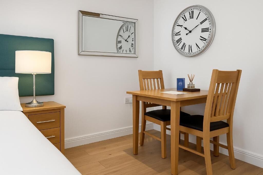A hotel room with a bed, a bedside table with a lamp, a wooden desk with two chairs, and two wall clocks.