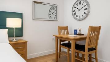 A hotel room with a bed, a bedside table with a lamp, a wooden desk with two chairs, and two wall clocks.