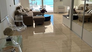 Uma sala de estar moderna com um sofá, cadeiras transparentes, uma mesa de centro, uma televisão e uma grande janela com vista para o mar.