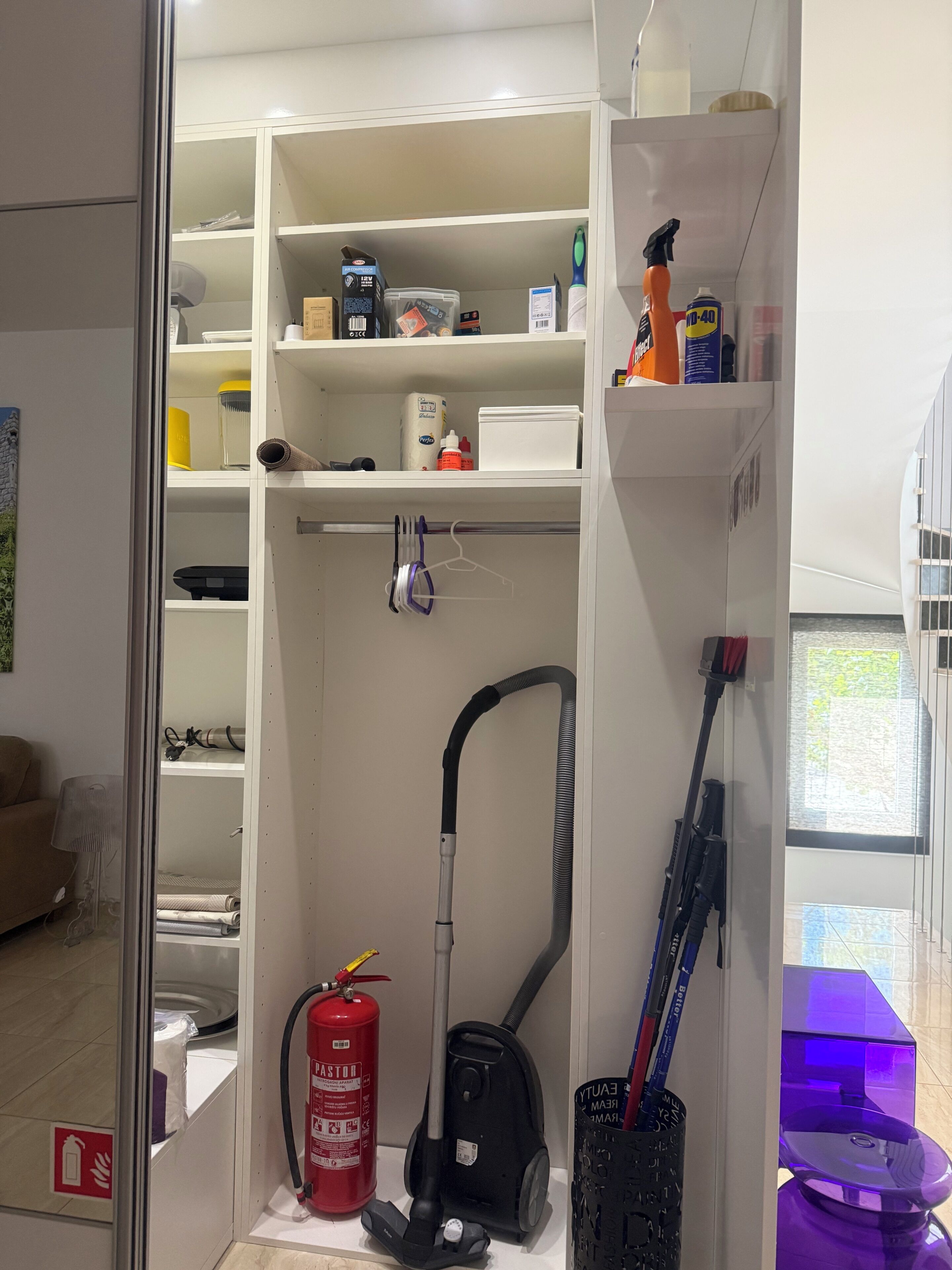 Um quarto com um armário branco, um aspirador de pó, um extintor de incêndio e uma janela com vista para o exterior.
