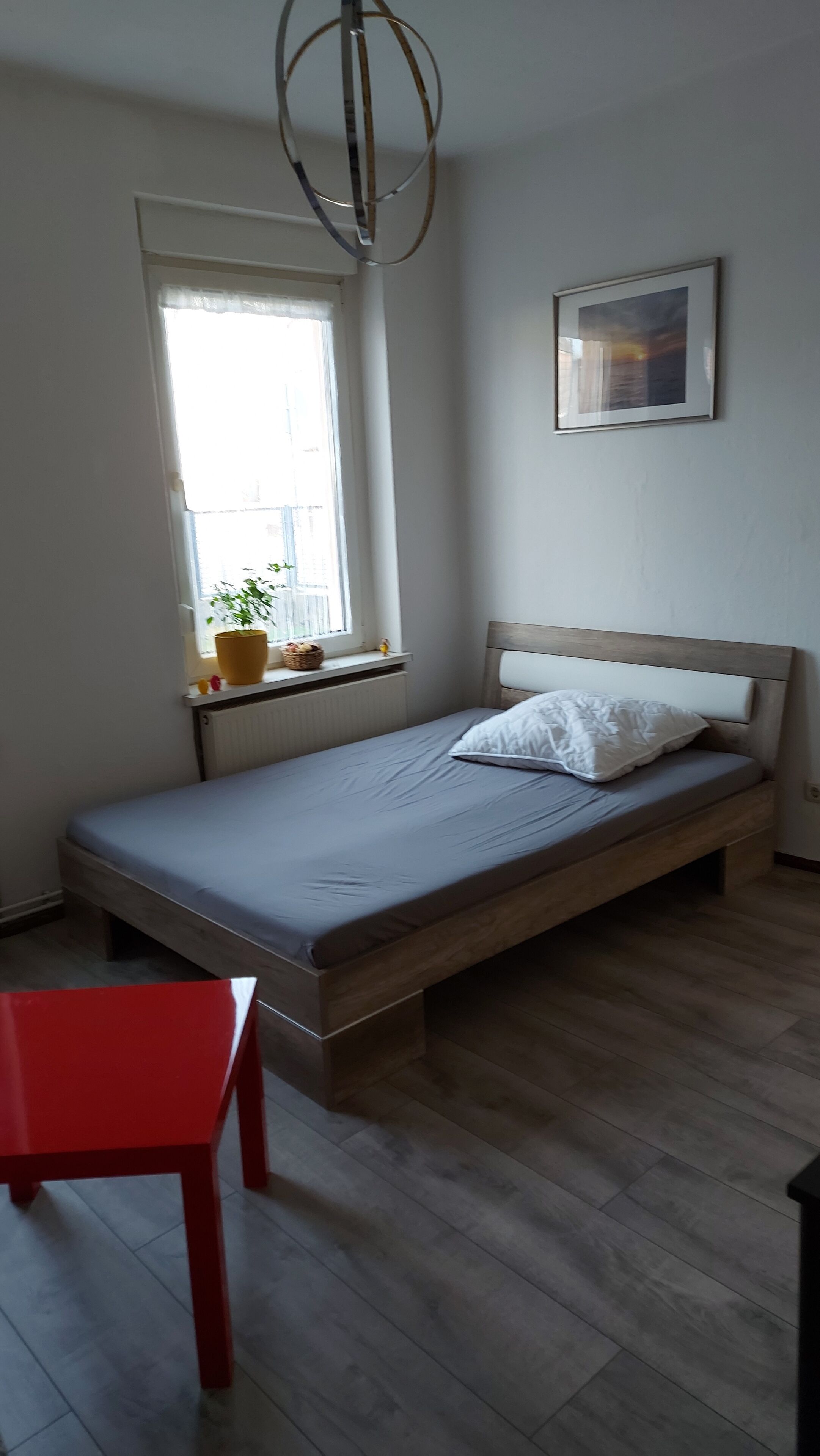 A bedroom with a bed, a window with a plant, a red chair, and a framed picture on the wall.