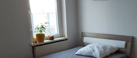 A bedroom with a bed, a window with a plant, a red chair, and a framed picture on the wall.