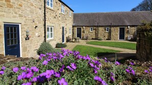 A stone building with a blue door, a small garden with purple flowers, and a grassy courtyard. - Chaff House – Dog‑Friendly Family Cottage on Burradon Farm near coast (Cramlington)