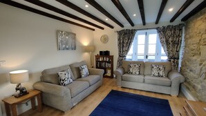 A living room with two sofas, a blue rug, wooden beams on the ceiling, a bookshelf, and a window with curtains. - Chaff House – Dog‑Friendly Family Cottage on Burradon Farm near coast (Cramlington)