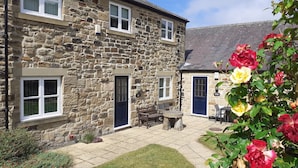 A stone building with a blue door, a small patio with seating, and a garden with flowers. - Chaff House – Dog‑Friendly Family Cottage on Burradon Farm near coast (Cramlington)