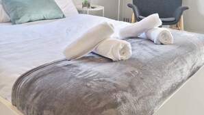 Un lit bien fait, avec une literie blanche et grise, une chaise grise, une petite table de chevet avec une lampe, et une fenêtre donnant sur des arbres.