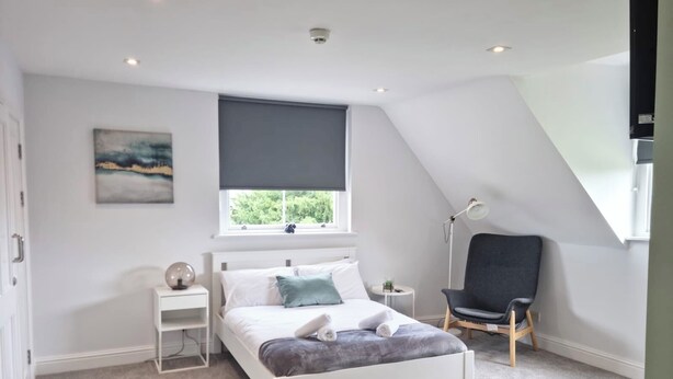 Une chambre avec un lit blanc, une chaise grise, une petite table de chevet, une lampe sur pied et une fenêtre donnant sur un paysage verdoyant.