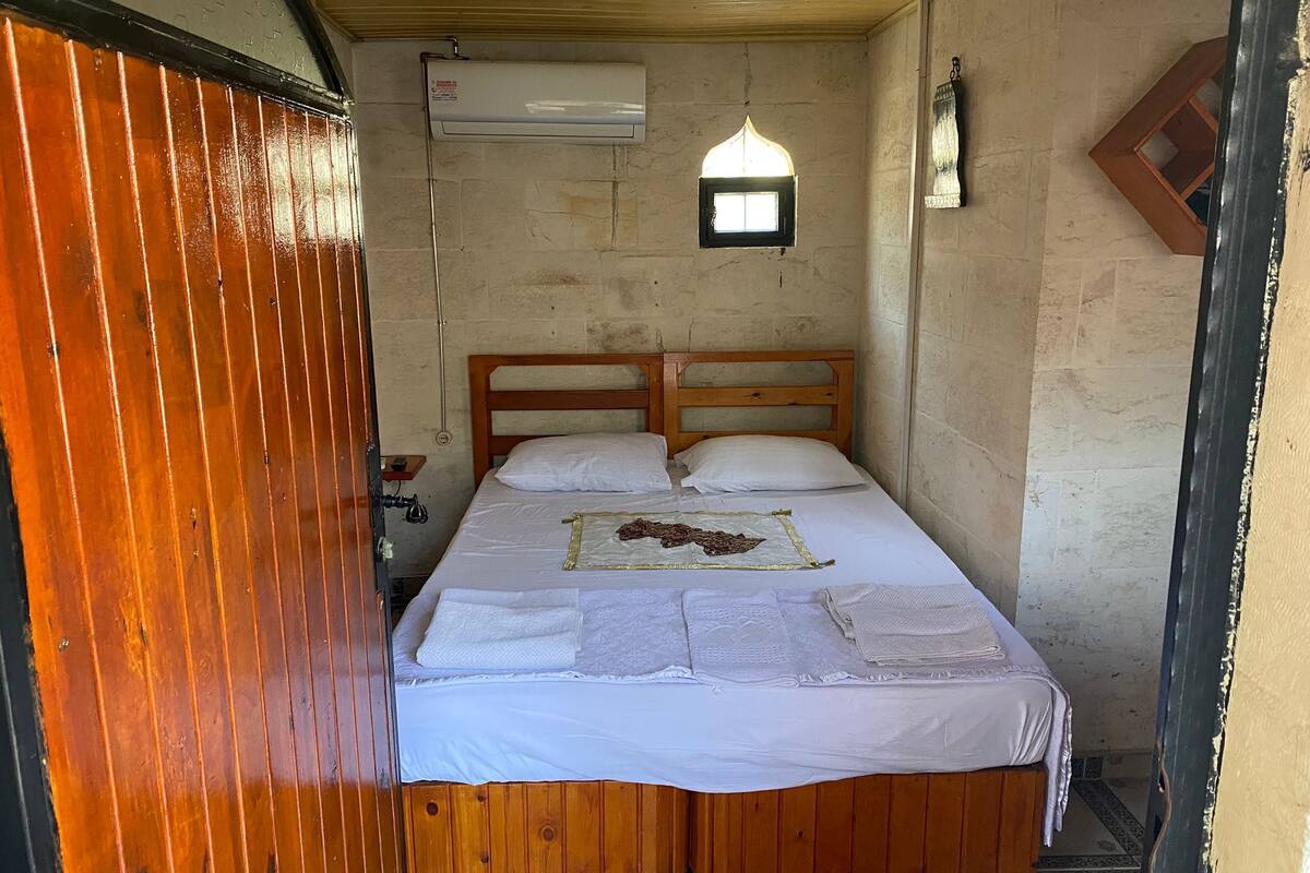 A small, rustic room with a wooden bed, a small window, and a wall-mounted air conditioner.