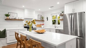 A modern kitchen with white cabinetry, a large island with bar stools, stainless steel appliances, and decorative plants.