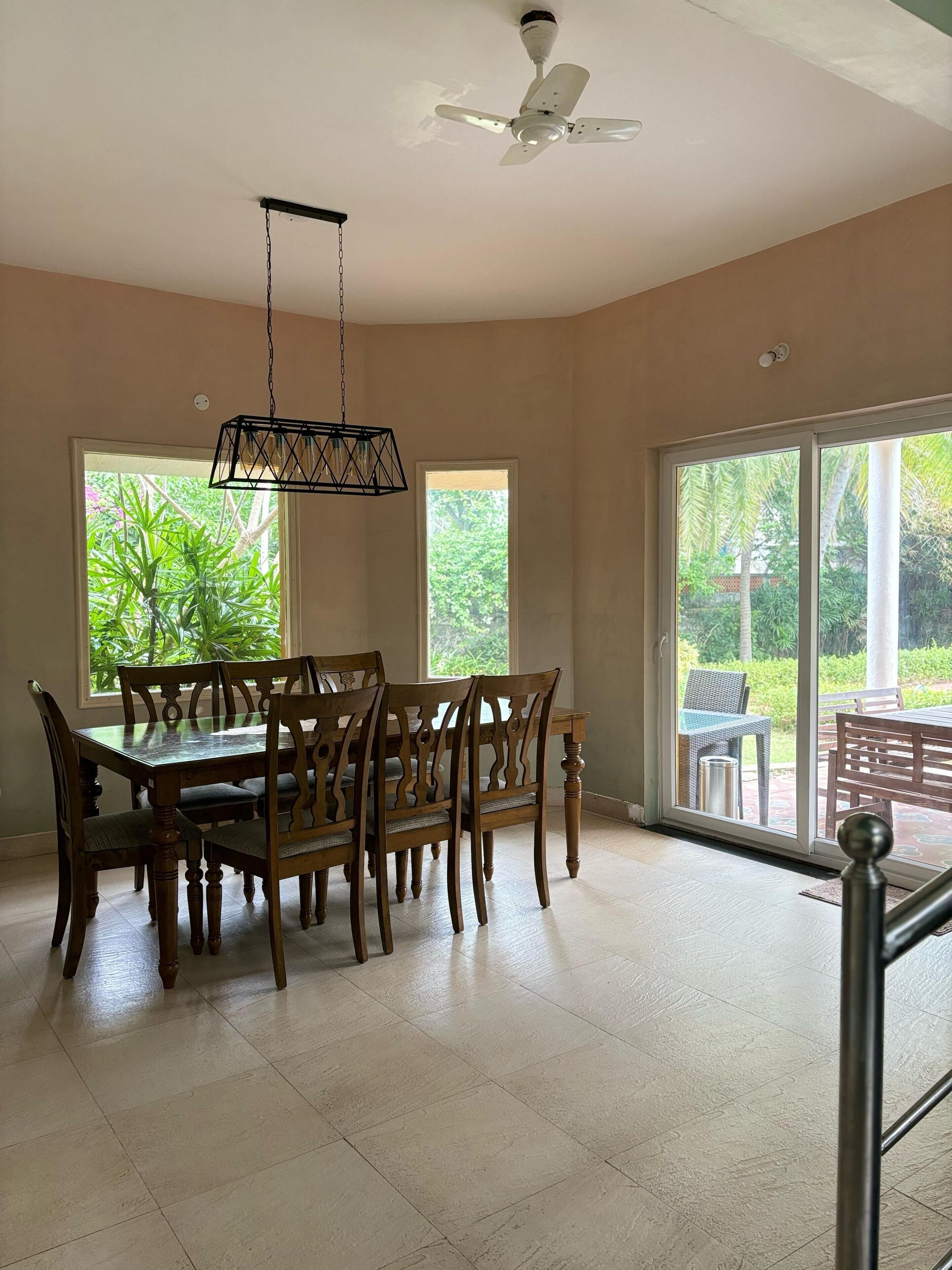 A dining area with a table and chairs, a ceiling fan, and large windows with a view of greenery.