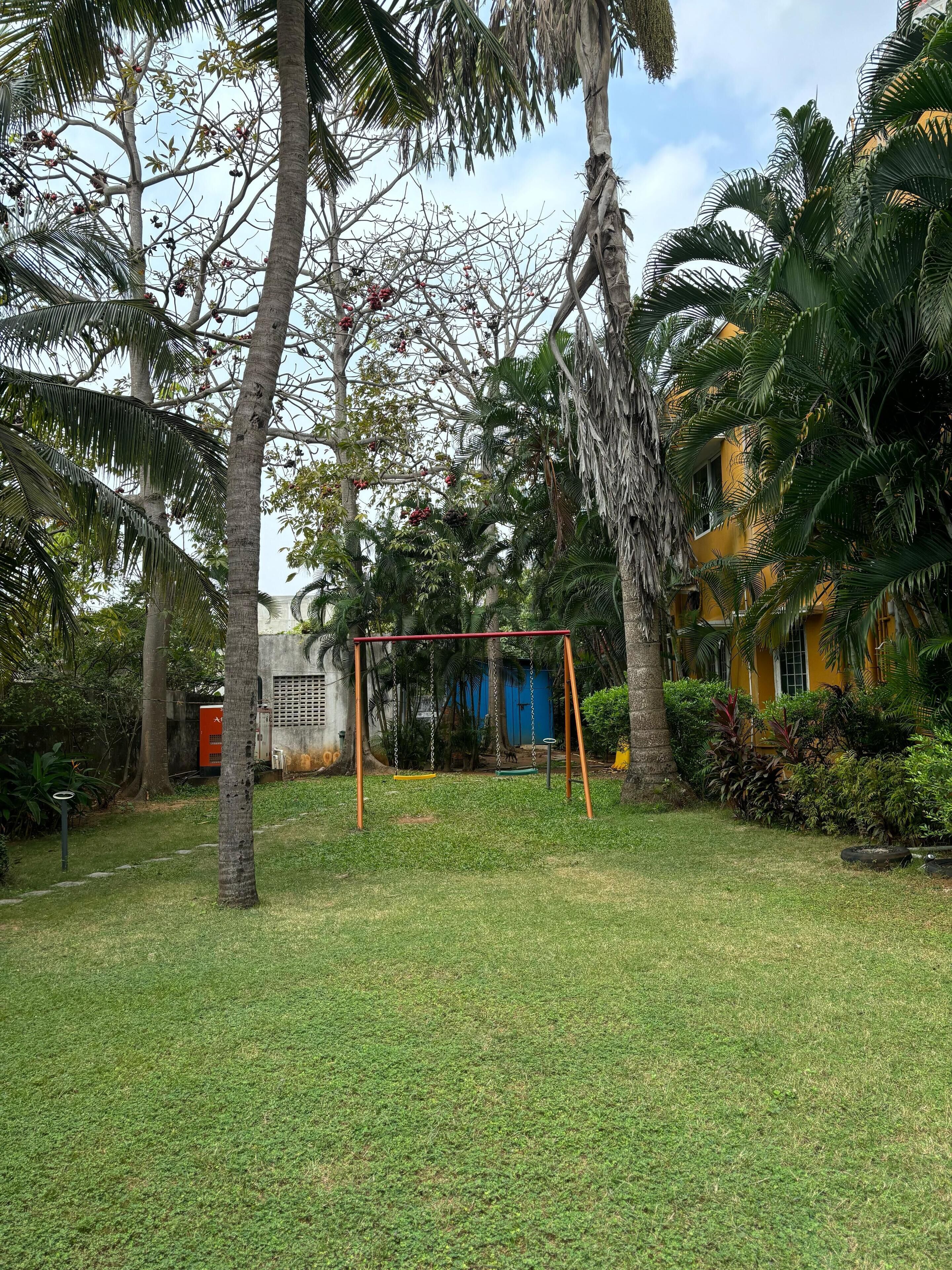 A grassy area with a swing set, palm trees, and a yellow building in the background.