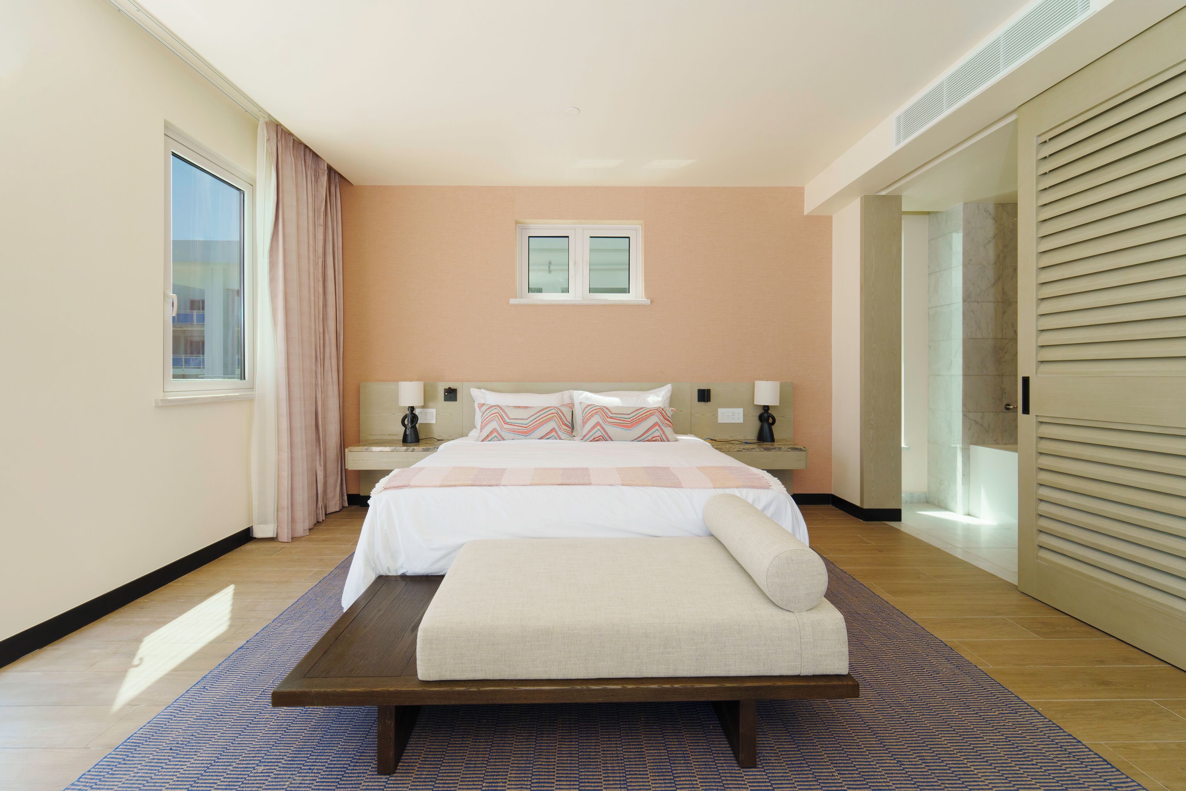 A hotel room with a large bed, a bench, a window with curtains, and a door with blinds.