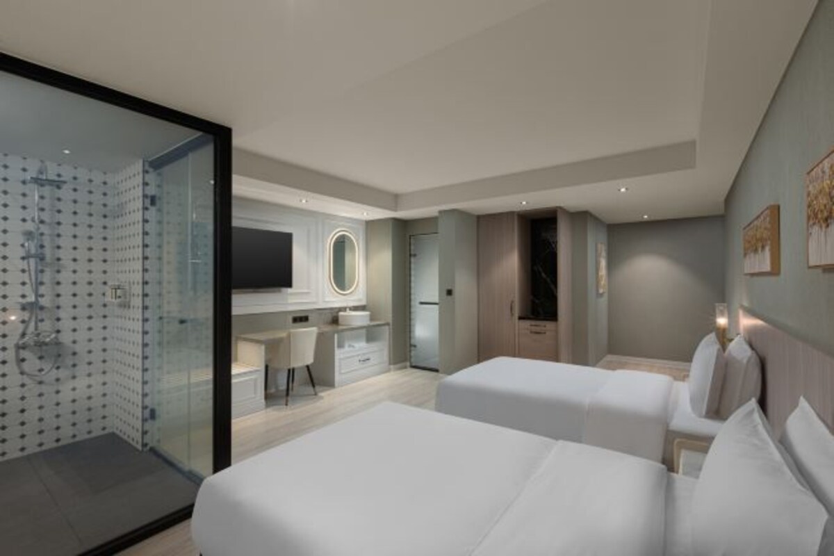 A modern hotel room with a glass shower enclosure, a vanity with a mirror, a desk, and two beds.