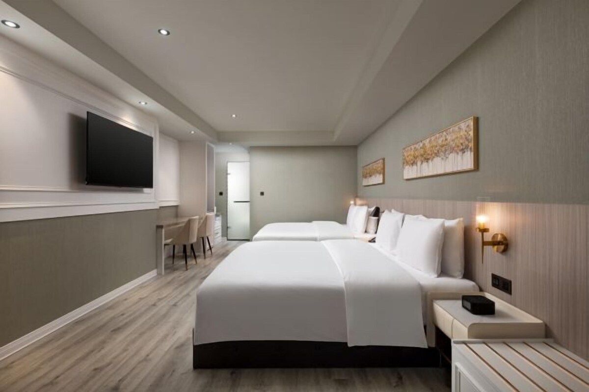 A hotel room with two beds, a flat-screen TV mounted on the wall, a desk with a chair, and a small table with a lamp.