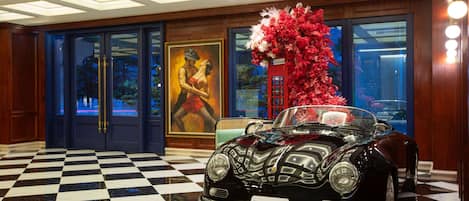 A classic car in a lobby with a checkered floor, a large floral arrangement, and a painting of a woman in a red dress.