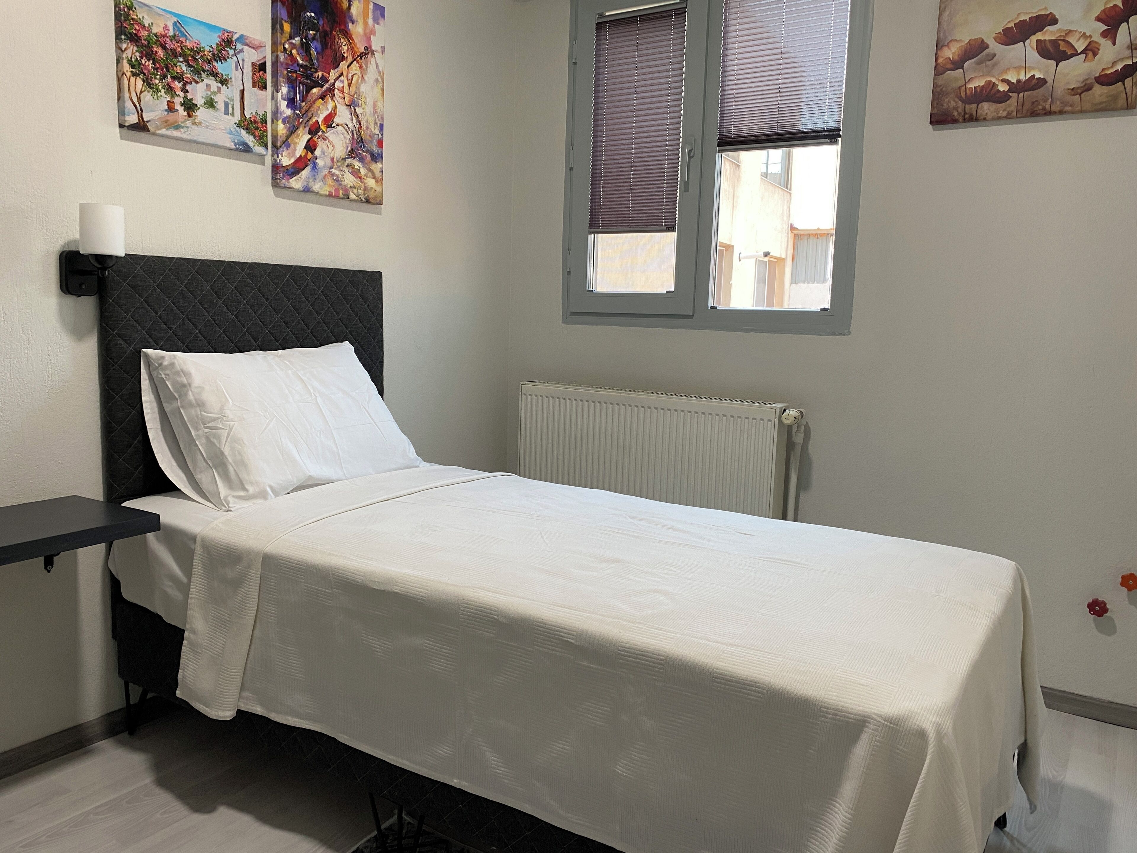 A bedroom with a bed, a bedside table, a radiator, and two windows with blinds.