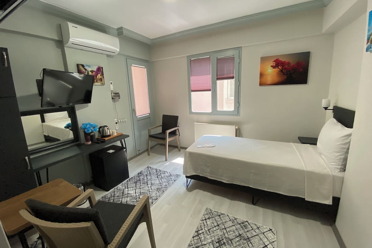 Ein kleines Hotelzimmer mit Bett, Schreibtisch mit Computer, Stuhl, Heizkörper und Fenster mit Jalousien.