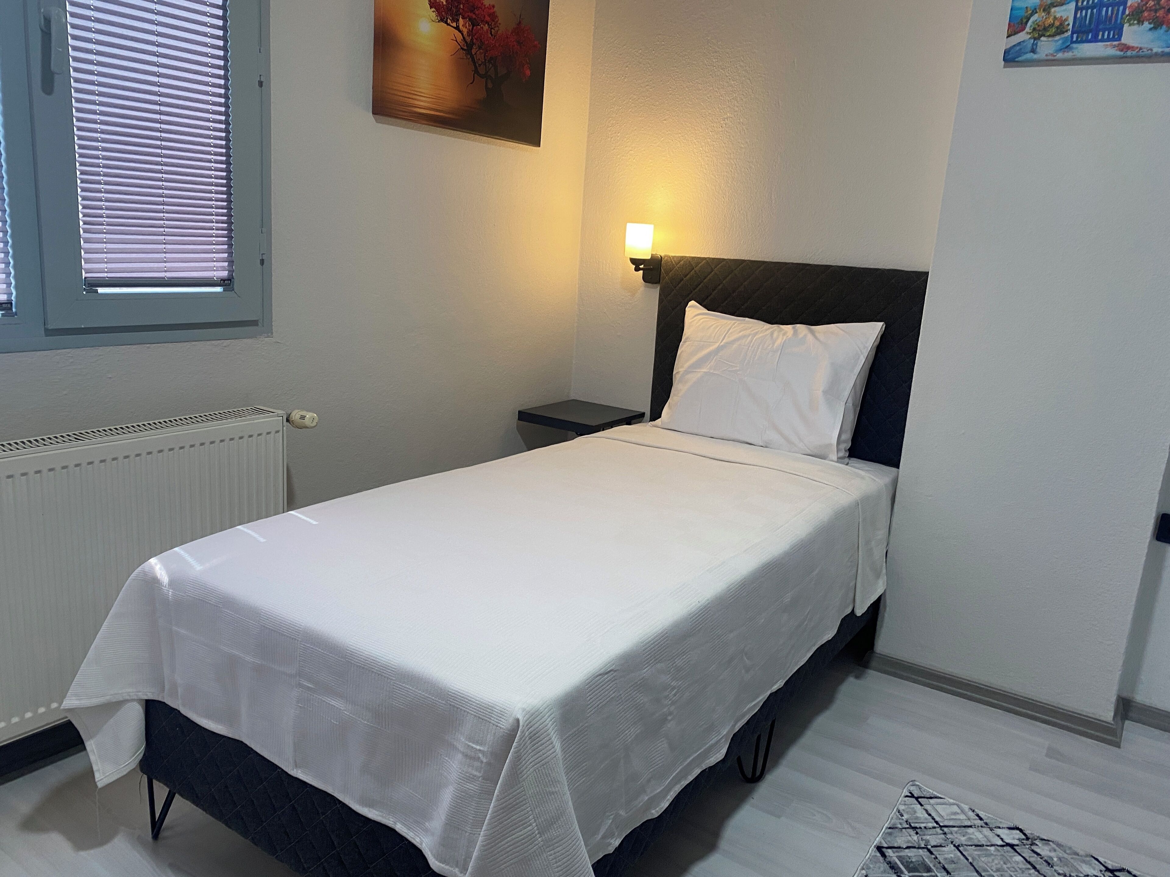 En pænt redt seng med hvid dyne, et natbord med lampe, en radiator og et vindue med persienner.