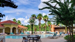 Een zwembadgedeelte met ligbedden en tafels, omgeven door palmbomen en een gebouw met een rood dak.